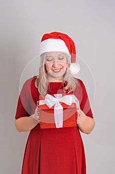 A beautiful adult woman in a Santa hat and a red dress poses on a gray background holding a hat pompom in her hands