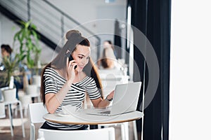 Beautiful adult woman in striped shirt is talking on the phone, holding a cup and smiling while working with a laptop in