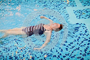 Beautiful adult woman in striped swimsuit swimming in blue pool on her back at resort. Sport activity health concept.