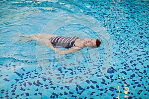 Beautiful adult woman in striped swimsuit swimming in blue pool on her back at resort. Sport activity health concept.