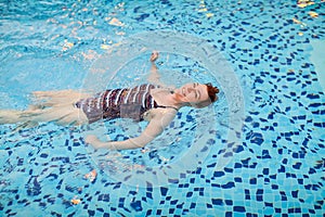 Beautiful adult woman in striped swimsuit swimming in blue pool on her back at resort. Sport activity health concept.