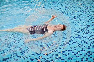 Beautiful adult woman in striped swimsuit swimming in blue pool on her back at resort. Sport activity health concept.