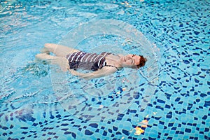 Beautiful adult woman in striped swimsuit swimming in blue pool on her back at resort. Sport activity health concept.