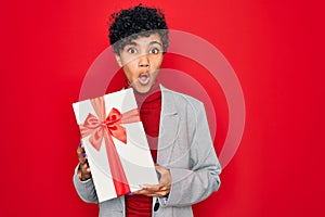 Beautiful african american afro business woman holding gift over isolated red background scared in shock with a surprise face,