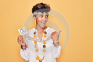 Beautiful african american afro hippie woman wearing sunglasses holding reminder peace sign pointing and showing with thumb up to