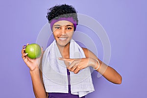 Beautiful african american afro sporty woman doing exercise wearing towel eating green apple very happy pointing with hand and