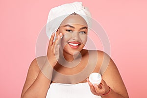 Beautiful african american body positive lady holding jar of cream, and applying moisturizer on cheek, pink background