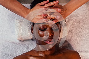 Beautiful african american female getting Ayurveda face massage by the white-skinned hands of man in spa salon.