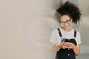 Beautiful African American girl in fashionable clothing, uses mobile phone for sending text messages, connected to wifi, checks