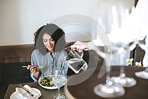 Beautiful African American girl sitting in restaurant and eating salad while waiter pouring water in glass Portrait of