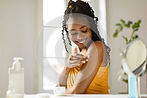 Beautiful african american lady applying cream on her shoulder, taking care of skin after bath in bedroom, free space