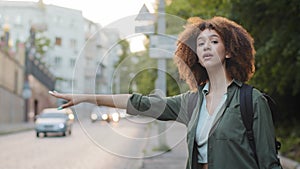 Beautiful African American millennial woman in casual wear waving down car, hitchhiking on roadside. Pretty young black