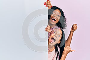 Beautiful african american mother and daughter holding blank empty banner pointing thumb up to the side smiling happy with open