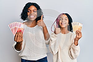 Beautiful african american mother and daughter holding norwegian krone banknotes smiling happy and positive, thumb up doing