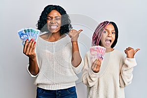 Beautiful african american mother and daughter holding south african rand banknotes pointing thumb up to the side smiling happy