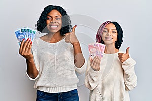 Beautiful african american mother and daughter holding south african rand banknotes smiling happy and positive, thumb up doing