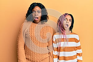 Beautiful african american mother and daughter wearing wool winter sweater in shock face, looking skeptical and sarcastic,