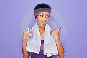 Beautiful african american sporty woman doing exercise wearing towel drinking glass of water pointing and showing with thumb up to
