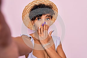 Beautiful african american tourist woman wearing bikini and hat making selfie by camera cover mouth with hand shocked with shame
