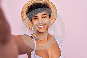 Beautiful african american tourist woman wearing bikini and hat making selfie by camera with a happy face standing and smiling