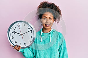 Beautiful african american woman with afro hair holding big clock looking positive and happy standing and smiling with a confident