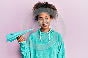 Beautiful african american woman with afro hair holding paper airplane looking positive and happy standing and smiling with a