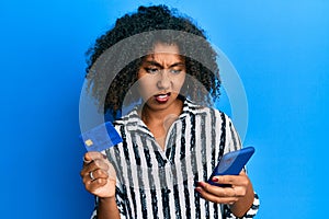 Beautiful african american woman with afro hair holding smartphone and credit card clueless and confused expression