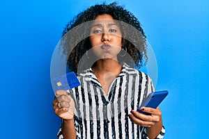 Beautiful african american woman with afro hair holding smartphone and credit card puffing cheeks with funny face