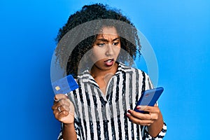 Beautiful african american woman with afro hair holding smartphone and credit card in shock face, looking skeptical and sarcastic,