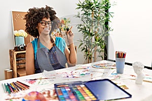 Beautiful african american woman with afro hair painting at art studio with a big smile on face, pointing with hand and finger to