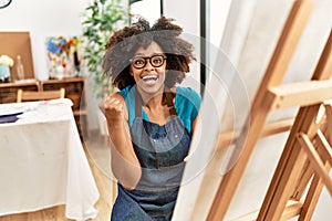 Beautiful african american woman with afro hair painting canvas at art studio screaming proud, celebrating victory and success