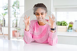 Beautiful african american woman with afro hair wearing casual pink sweater smiling funny doing claw gesture as cat, aggressive