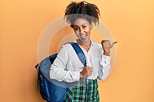 Beautiful african american woman with afro hair wearing student backpack pointing thumb up to the side smiling happy with open