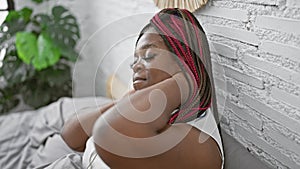 Beautiful african american woman with braids suffering from cervical pain, sitting unhappily on cozy bed in sunny bedroom, a