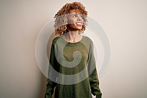 Beautiful african american woman with curly hair wearing casual sweater over white background looking away to side with smile on