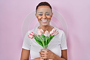 Beautiful african american woman holding bouquet of pink tulips flowers smiling with a happy and cool smile on face