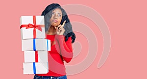 Beautiful african american woman holding presents serious face thinking about question with hand on chin, thoughtful about