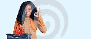 Beautiful african american woman holding supermarket shopping basket doing ok sign with fingers, smiling friendly gesturing