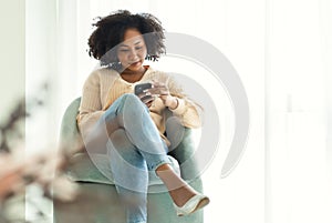 Beautiful African american woman sitting in comfy armchair, feeling relax using smartphone device at home. Happy black lady enjoy