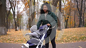 Beautiful African American young mother looking at camera smiling with cute curios boy sitting in baby stroller outdoors