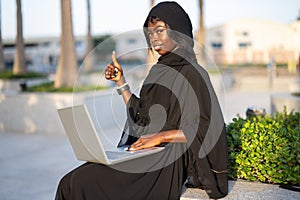 Beautiful African College female student in abaya with laptop sitting outdoors and showing thumb up.