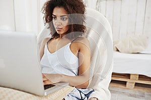 Beautiful african girl in sleepwear looking at laptop sitting in chair at home. Copy space.