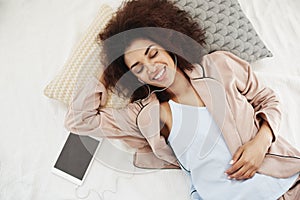 Beautiful african girl in sleepwear smiling listening to music in headphones lying on bed. Closed eyes. From above.