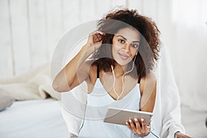 Beautiful african girl in sleepwear smiling looking at camera listening to music in headphones sitting on bed.