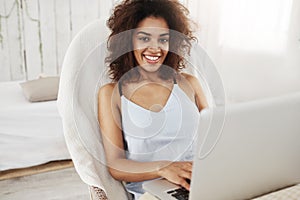 Beautiful african girl in sleepwear smiling looking at camera sitting in chair with laptop at home. Copy space.