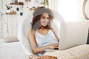 Beautiful african girl in sleepwear smiling looking at laptop sitting in chair at home. Copy space.