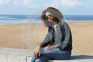 Beautiful african woman in a denim jacket and sunglasses is resting while sitting on the beach