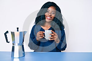 Beautiful african woman drinking a coffee from italian coffee maker looking positive and happy standing and smiling with a