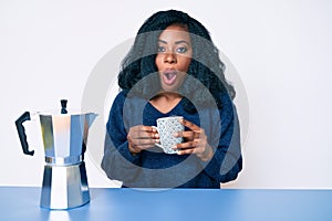 Beautiful african woman drinking a coffee from italian coffee maker scared and amazed with open mouth for surprise, disbelief face