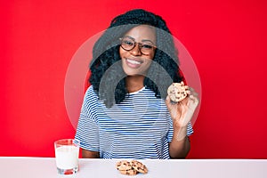 Beautiful african woman drinking a glass of fresh milk with chocolate cookies looking positive and happy standing and smiling with